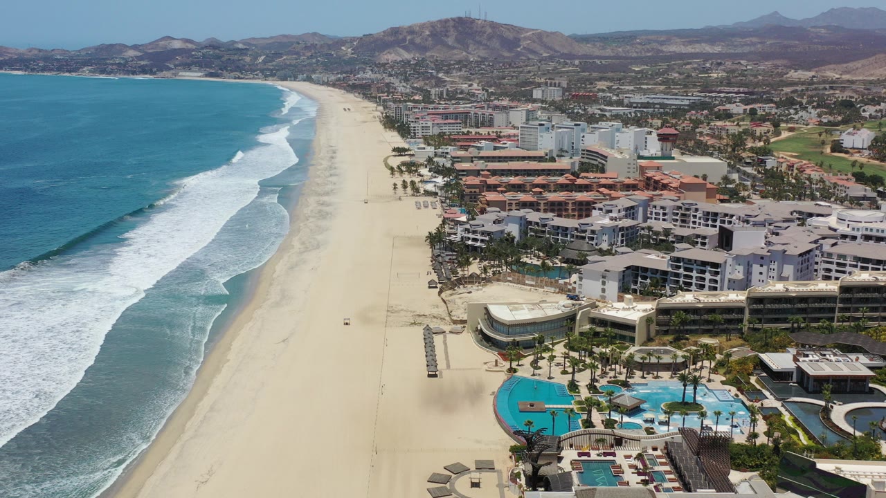 Aerial view of ocean surf beside a long sandy beach and resort buildings.