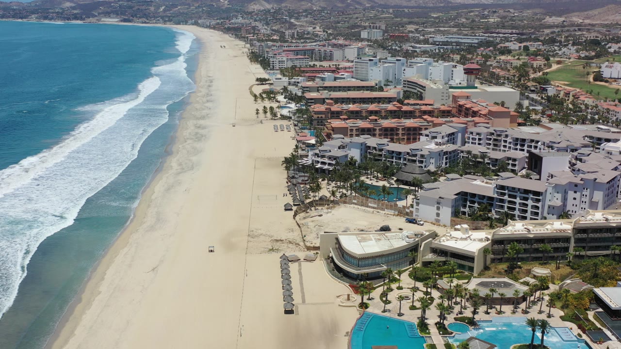 Aerial view in Los Cabos, Mexico, showing turquoise ocean, white surf, a wide sandy beach, beachfront hotels and condos, pools, palms, inland streets, and no visible people.