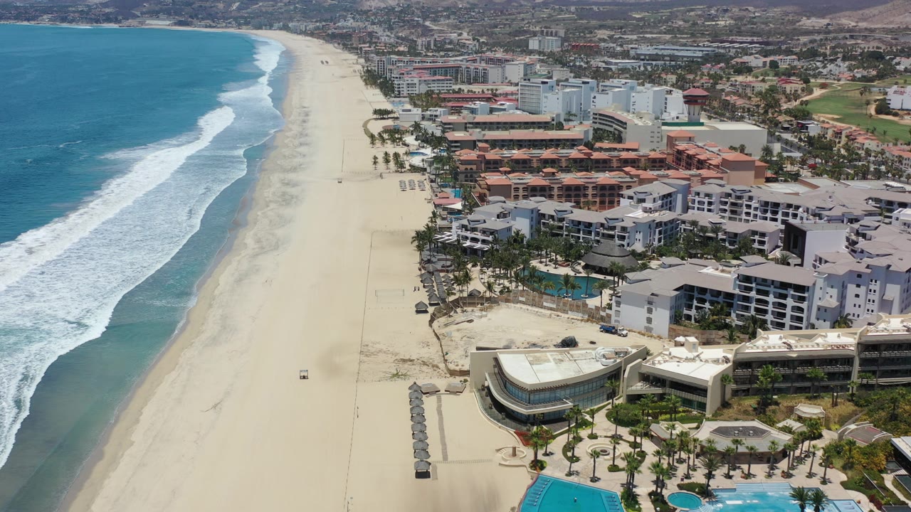 Drone view of ocean waves, beach, resort pools, and condos.