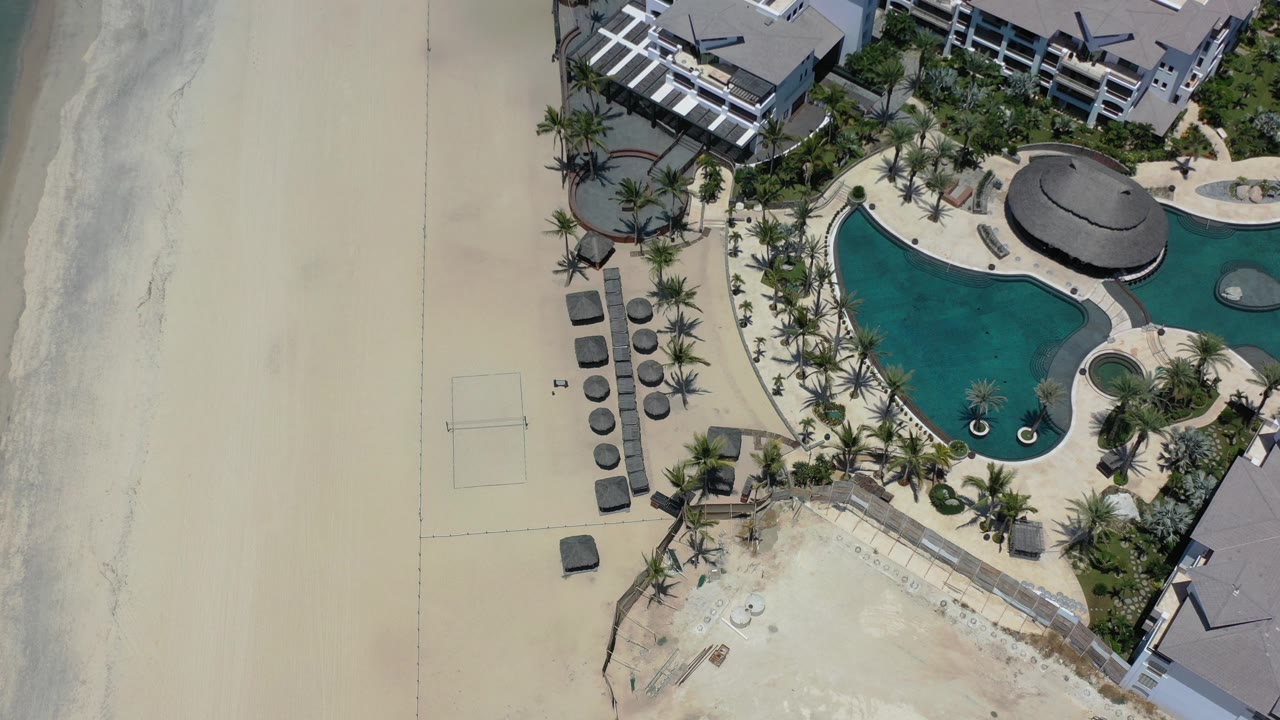 Top-down aerial frame in Los Cabos showing pale beach sand, a surf edge, palm landscaping, and a turquoise hotel pool with no visible people.