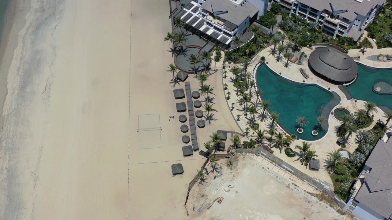 Overhead Los Cabos view with the hotel pool centered to the right, open sand to the left, surf along the beach edge, and no visible people.