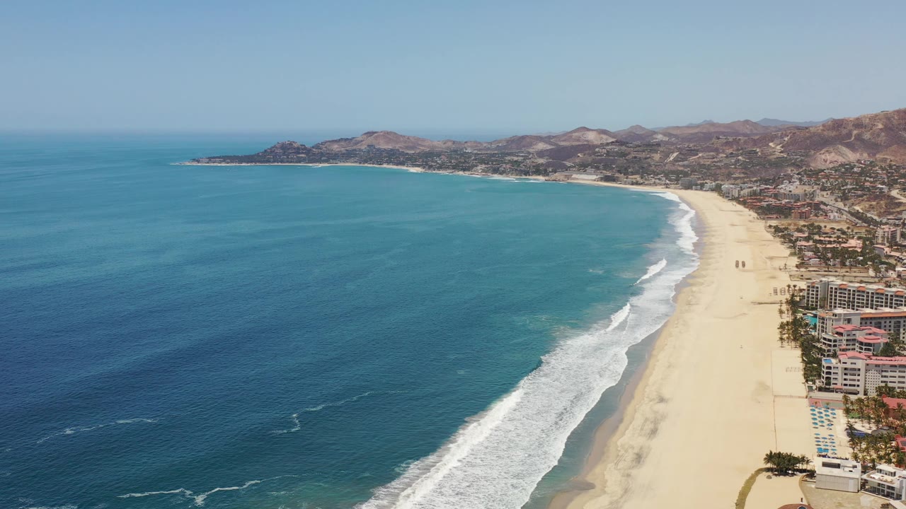 Aerial view in Los Cabos showing turquoise ocean across the left half, a curving surf band beside pale sand, resort-like buildings near the lower right edge, distant hills, and no visible people.