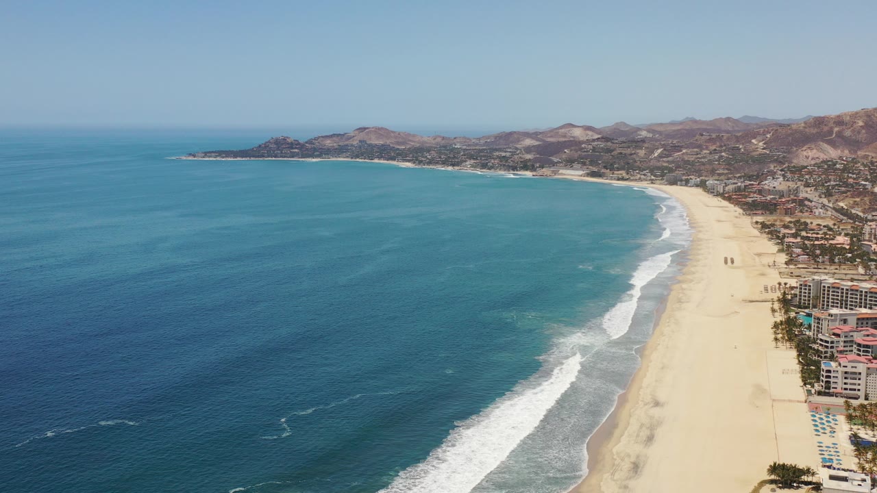 Opening aerial frame over Los Cabos, Mexico, with deep blue ocean filling the left side, a long sandy beach curving right, white surf lines, distant hills, and no visible people.
