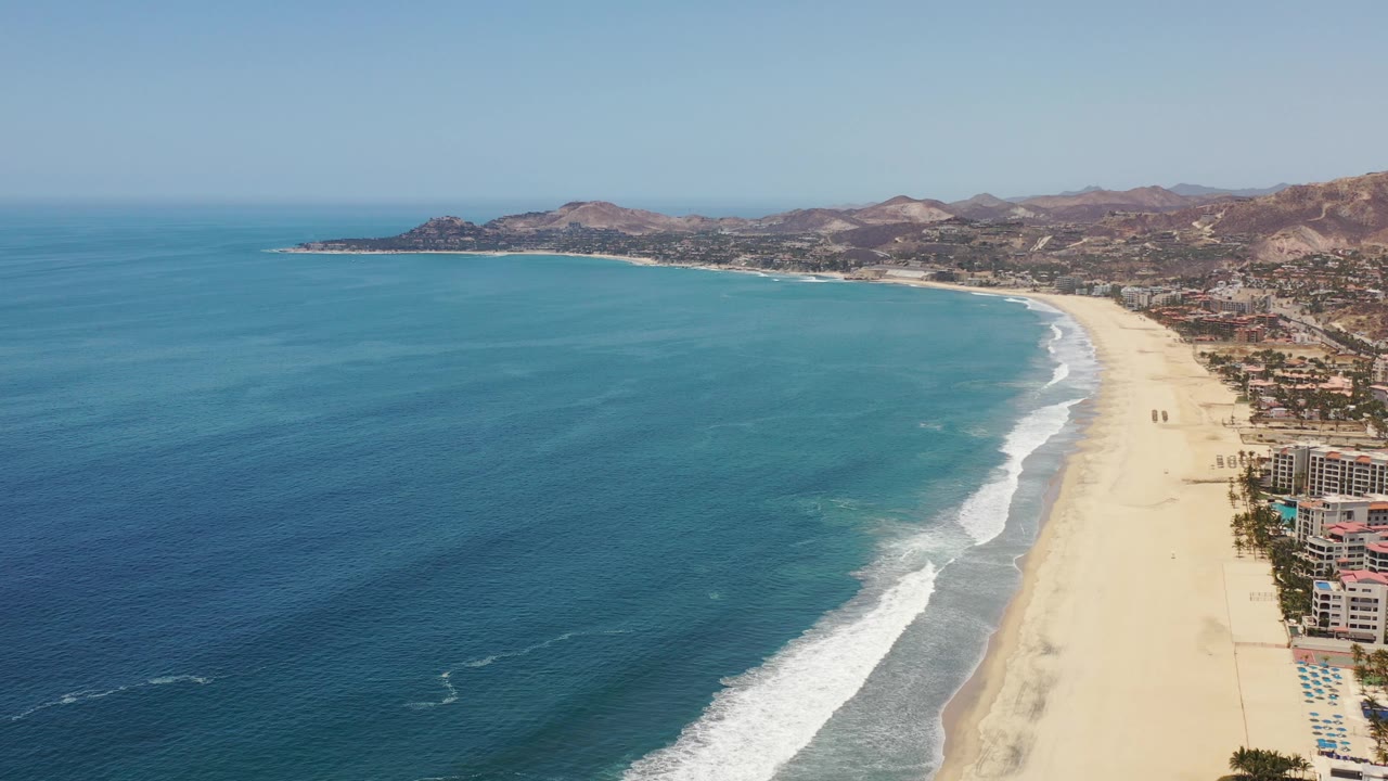 Aerial beach view with curved surf, turquoise shallows, ocean, buildings, and hills.
