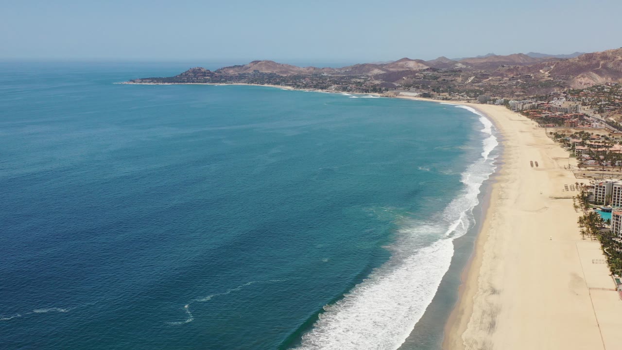 Aerial view of blue ocean, curved sandy beach, surf, city edge, and hills.