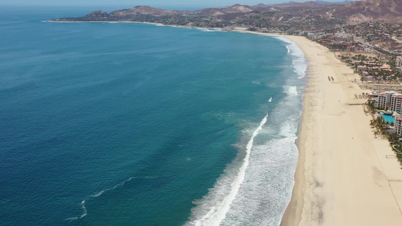 Drone view of surf rolling onto a broad beach beside coastal buildings.