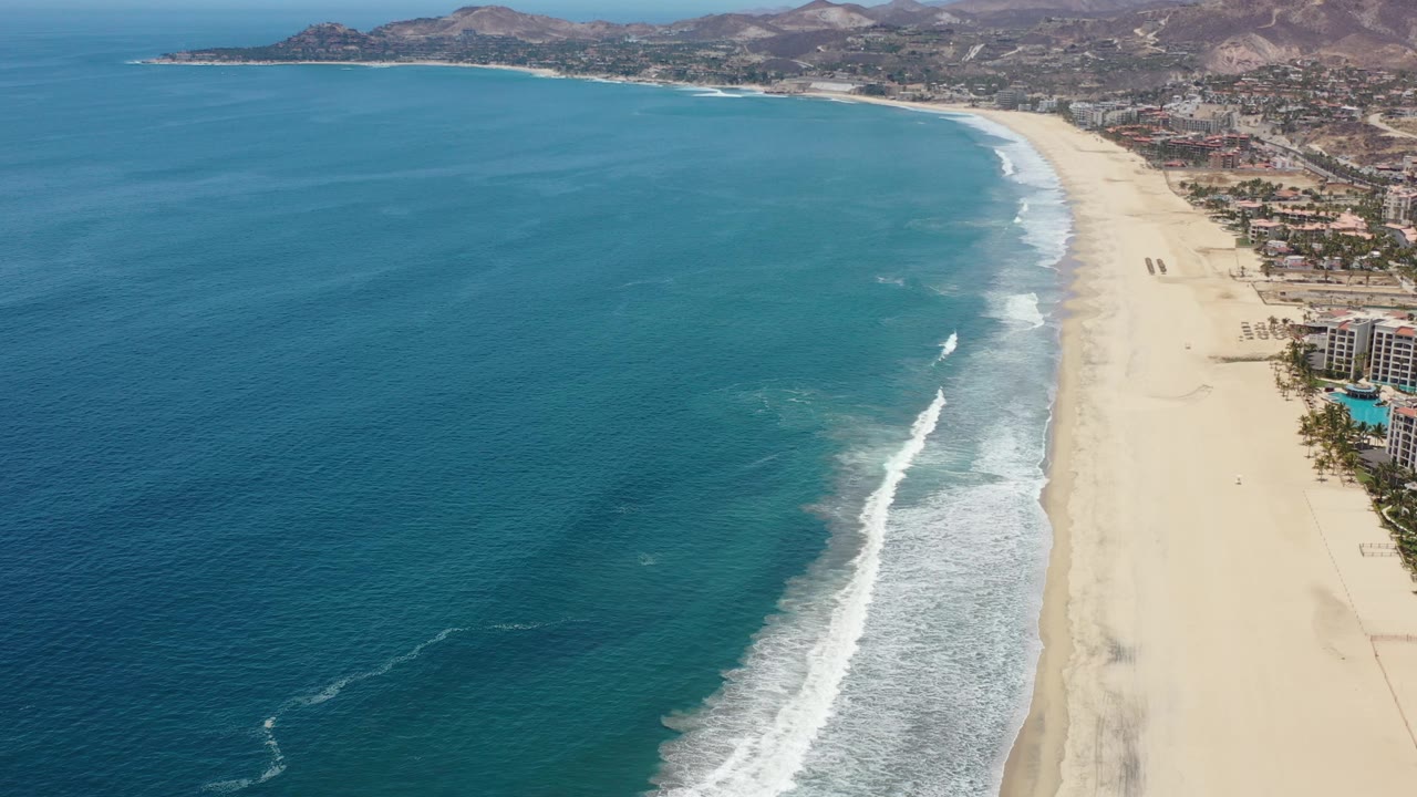 Aerial view of Los Cabos, Mexico, with open blue water dominating the left side, a long surf break near the beach, resort frontage to the right, and a hillside developed area in the background.