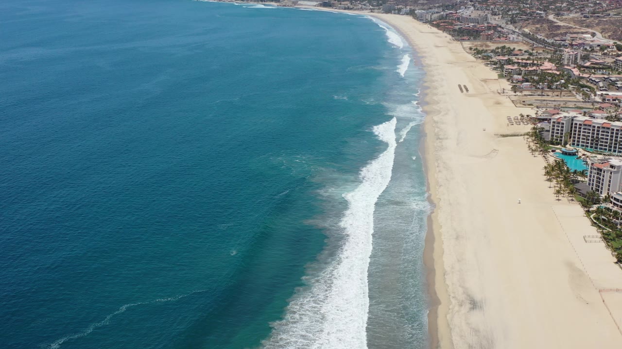 Opening aerial frame over the Los Cabos, Mexico coast, with blue ocean filling the left side, white surf meeting a long sand beach, shoreline developments at right, and no visible people.
