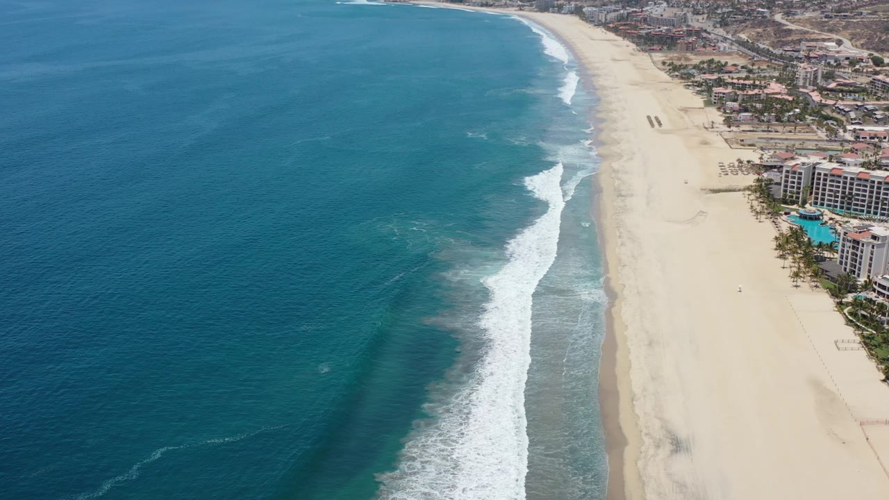 Oblique drone-style view of ocean waves beside a wide developed beach.
