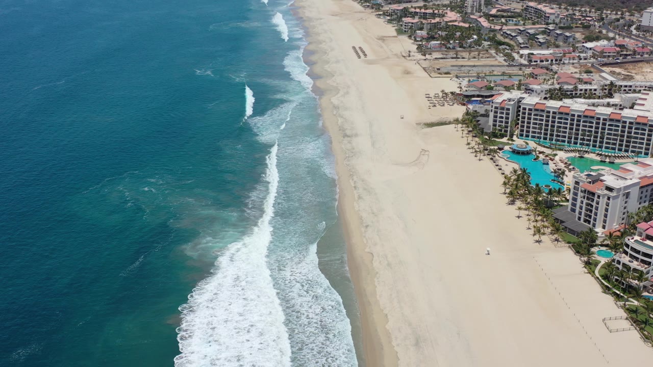 Aerial view over a Los Cabos beach with turquoise water on the left, white surf reaching pale sand, resort buildings and pools on the right, and no visible people.