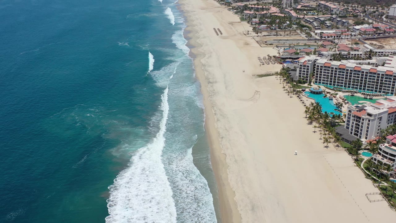 Drone-style view along the Los Cabos shoreline as a white wave line runs beside the sand and a large beachfront complex with blue pools sits to the right, no visible people.