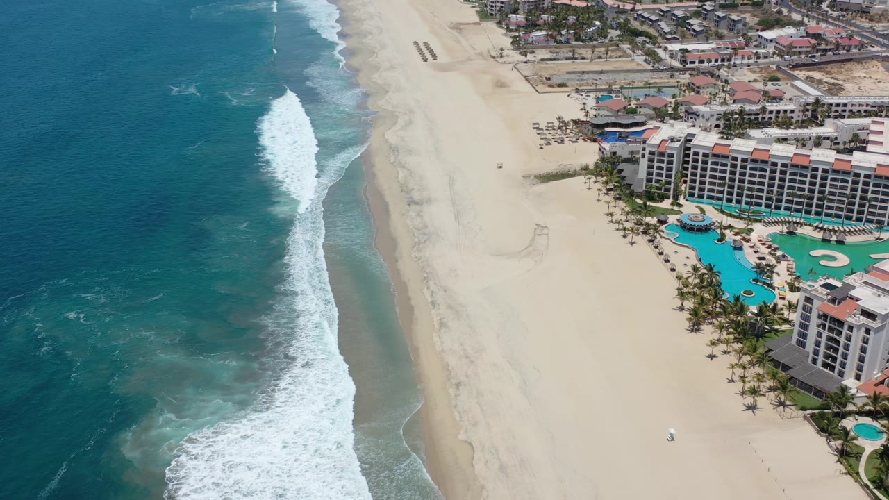 A second high aerial frame in Los Cabos shows the surf line slightly farther along the beach, with empty sand between the ocean and the resort pool area, and no visible people.