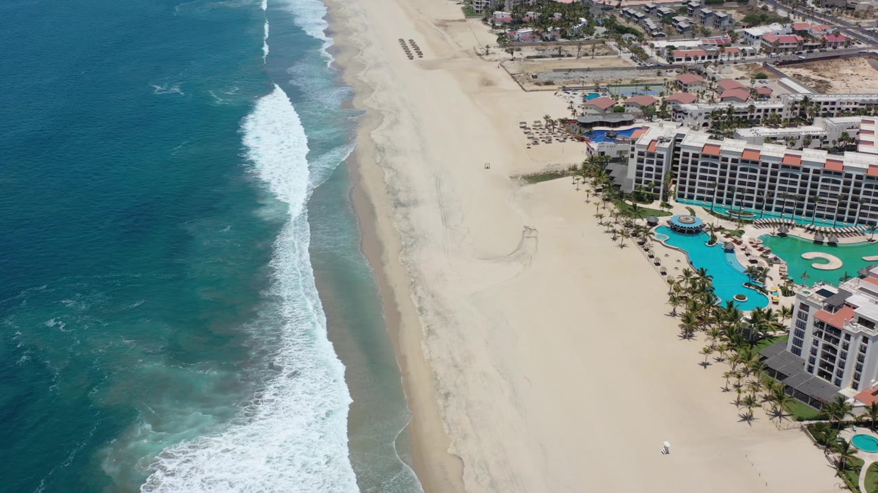 High above the Los Cabos shoreline, the frame places the ocean and breaking waves on the left, open sand in the center, and resort pools and hotel balconies at the right, with no visible people.