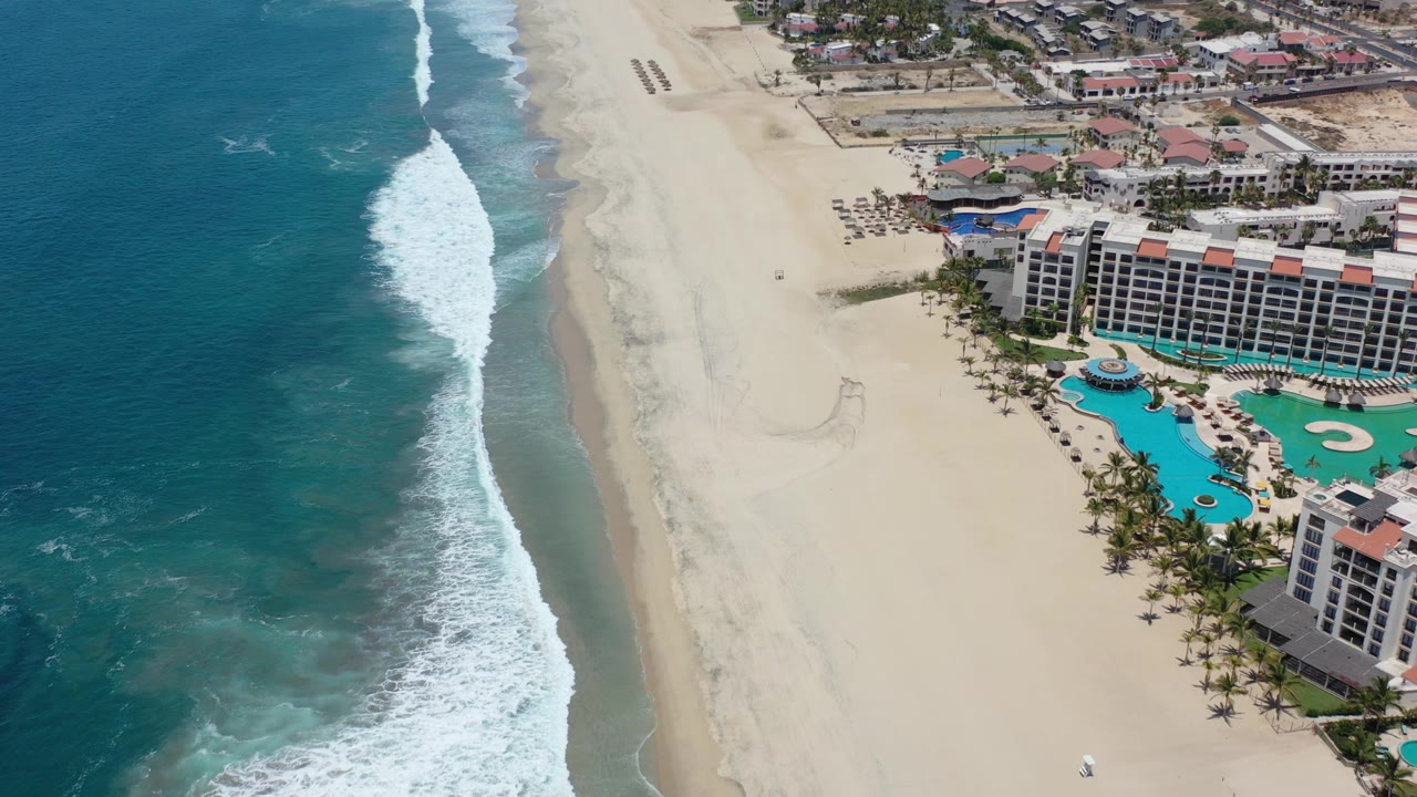 Aerial coastline with breaking surf, broad sand, and resort pool decks.