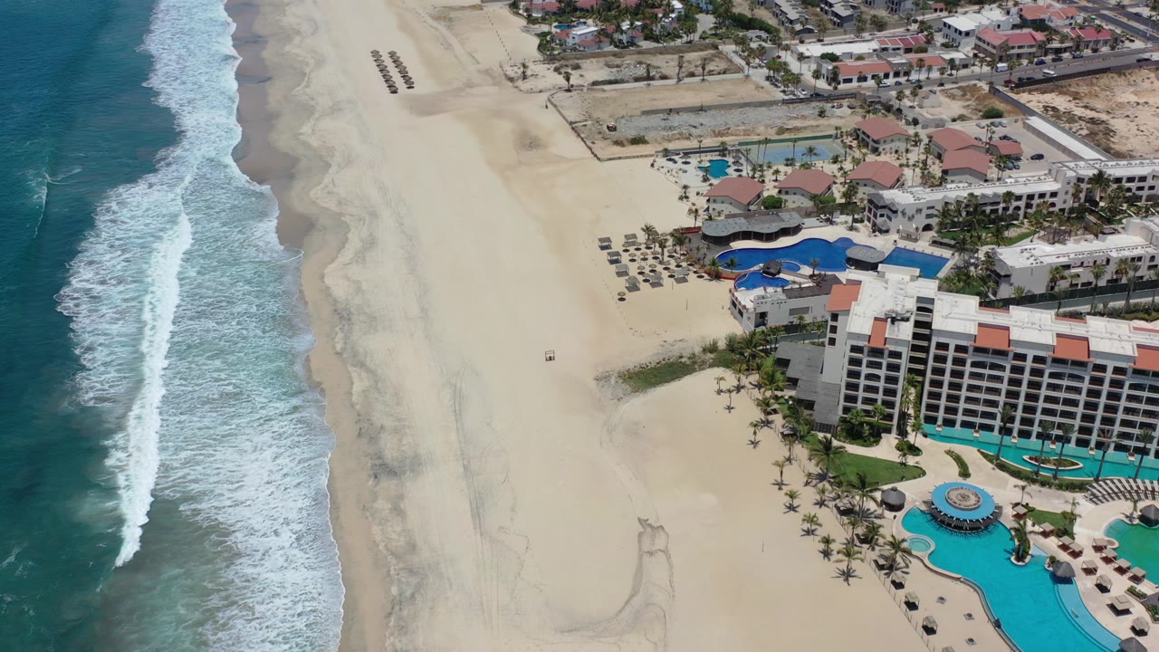 A high-angle aerial frame over Los Cabos, Mexico shows white surf on the left, wide pale sand in the center, resort pools and buildings on the right, and no visible people.