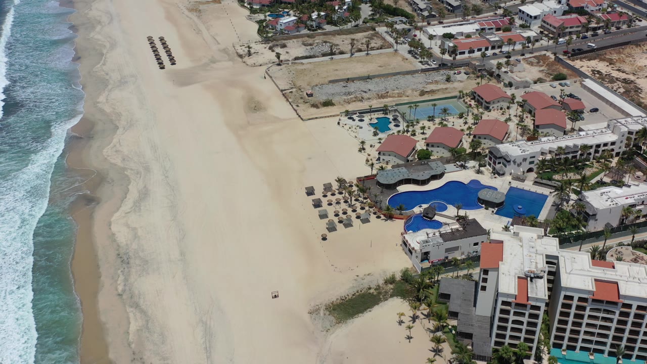 Los Cabos aerial view shows the ocean shifted more to the right side, with a wide beach, resort pool complex, rows of beach umbrellas, and houses beyond the shoreline road.