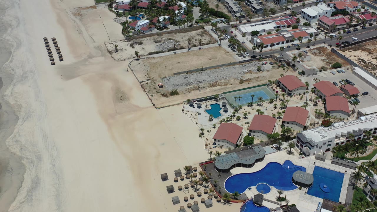 Aerial frame over Los Cabos resort grounds with a broad empty sand area at left, blue pools at bottom, red-roofed buildings, a tennis court, and no visible people.