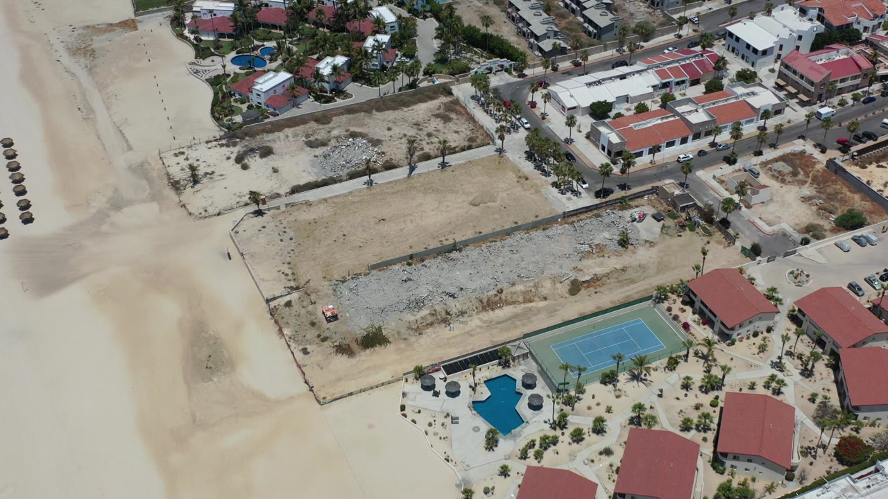 High aerial view in Los Cabos, Mexico showing pale beach sand, a fenced construction lot, red-roof buildings, a blue tennis court, a small pool, roads, parked cars, and no visible people.