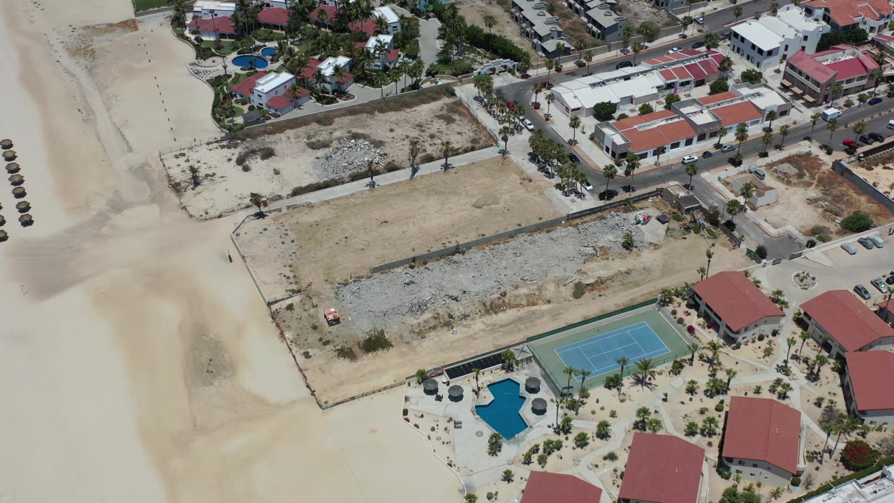 Aerial frame in Los Cabos, Mexico with the tennis court and pool lower in view, unfinished lots across the center, beach sand along the left and lower areas, and no visible people.