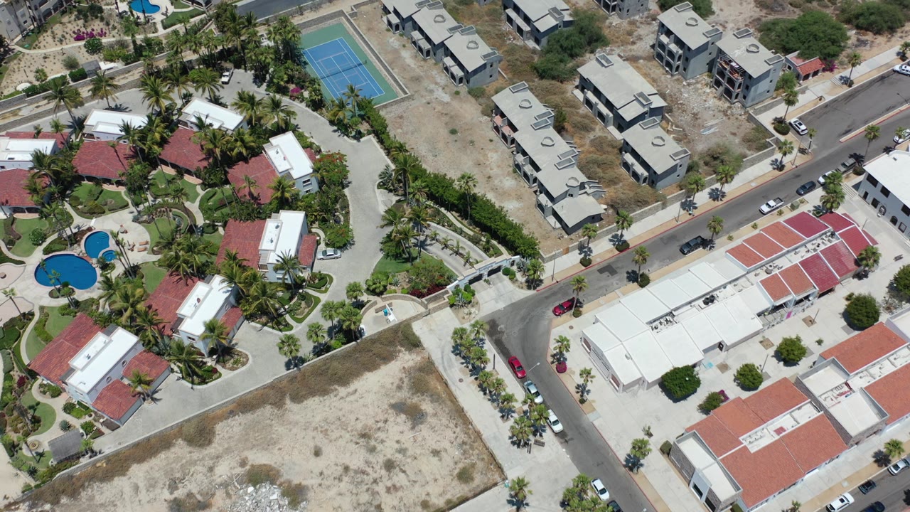 Aerial view of beachfront homes, pools, tennis court, roads, palms, and sandy lots.