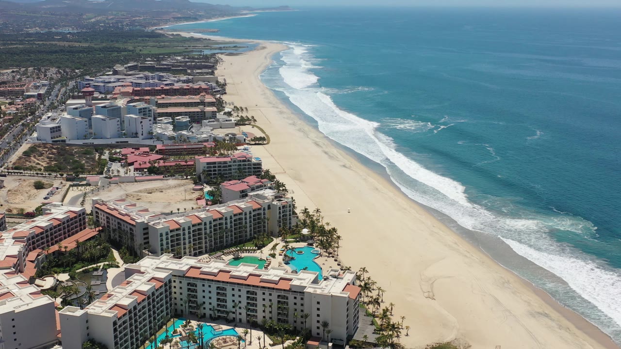 High view of the Los Cabos coastline with red-roof and light-colored resort buildings in the lower frame, pale sand through the center, blue water to the right, and no visible people.