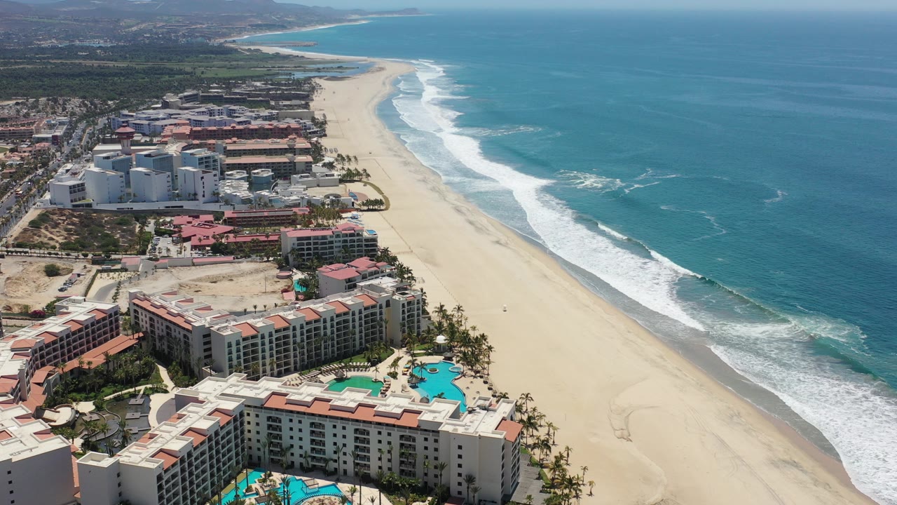 Aerial view over Los Cabos, Mexico beachfront resorts, turquoise pools, a long sandy shoreline, and white surf breaking along blue water, with no visible people.