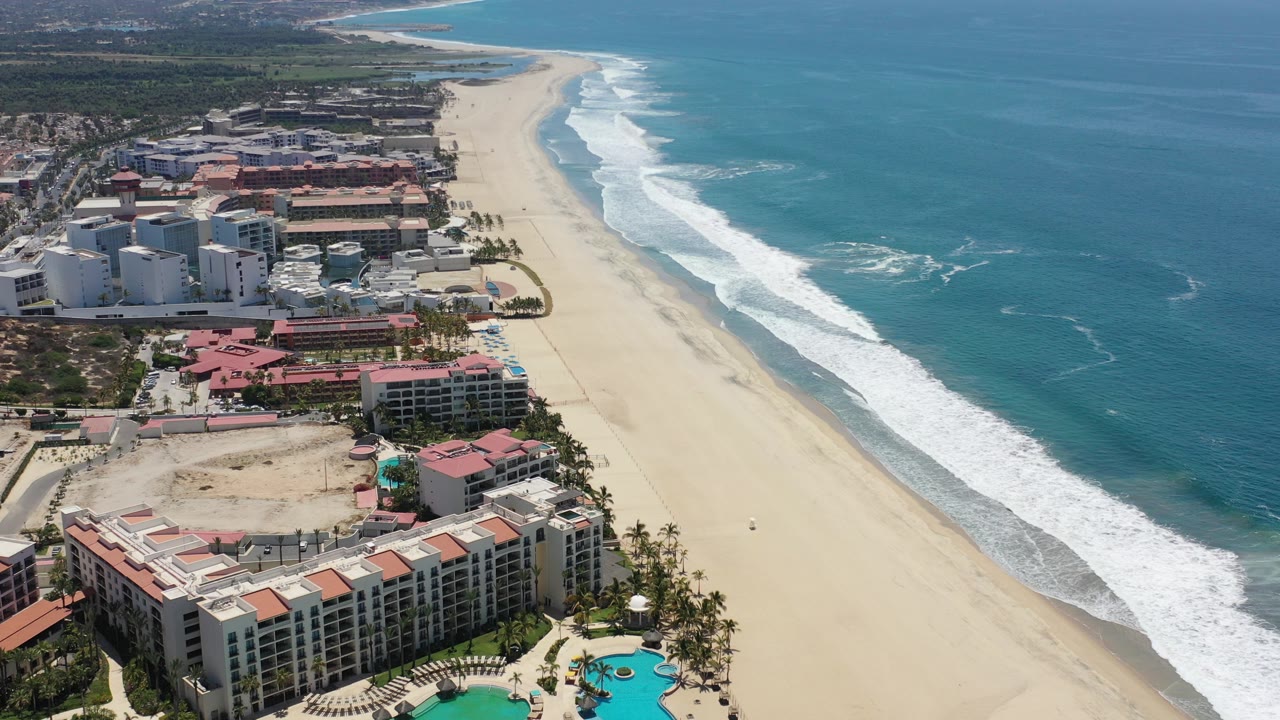 Opening frame of a Los Cabos, Mexico shoreline from above, with hotel buildings at left, pale sand through the center, blue ocean at right, white surf bands, and no visible people.