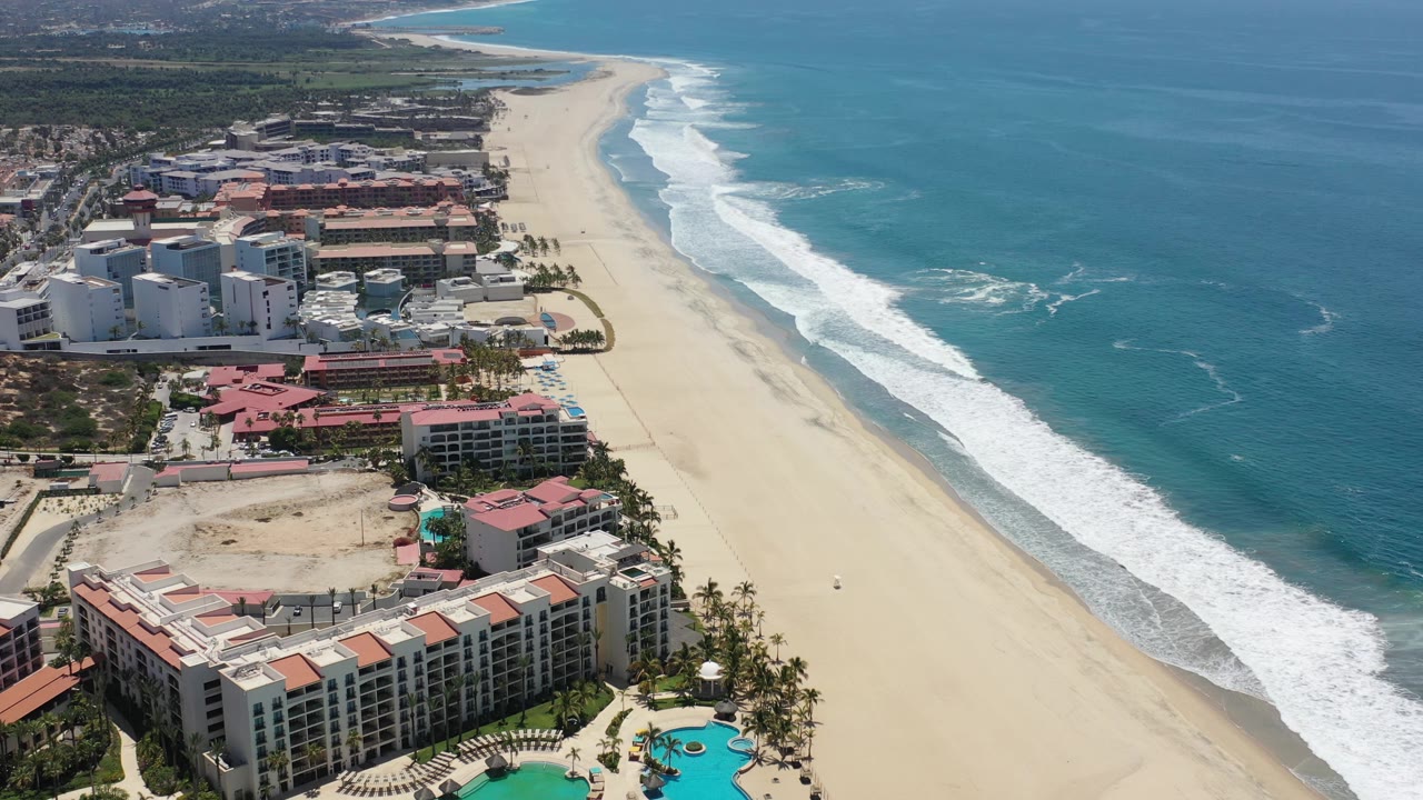 A slightly later Los Cabos aerial frame keeps the hotel blocks in the lower left while the long empty beach and white wave line extend toward the upper right, with no visible people.