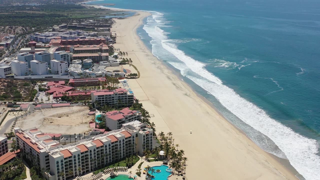 Oblique aerial coastline with hotels, beach, waves, and open sea.