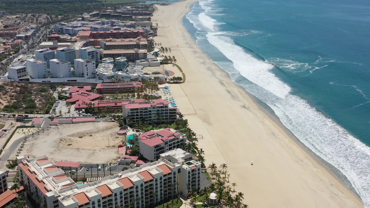 Overhead view in Los Cabos, Mexico showing resort buildings along a broad sandy beach with white surf on the right and no visible people.
