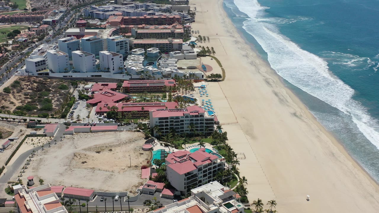 Aerial view over Los Cabos, Mexico resort buildings with red roofs, white hotel towers, pools, palms, broad sand, blue surf, and no visible people.