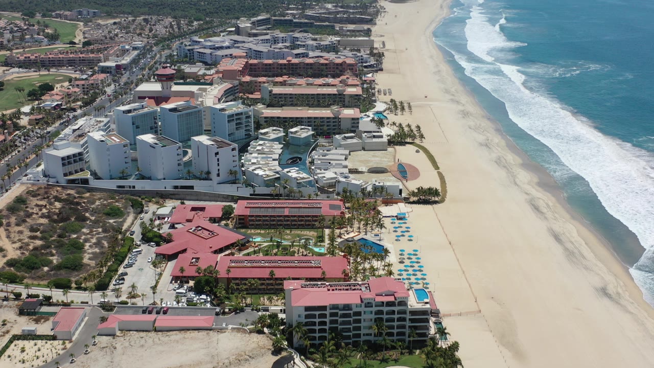 Aerial view in Los Cabos, Mexico, showing white beachfront buildings, red-roofed property areas, pale sand, blue ocean, white surf, and no visible people.