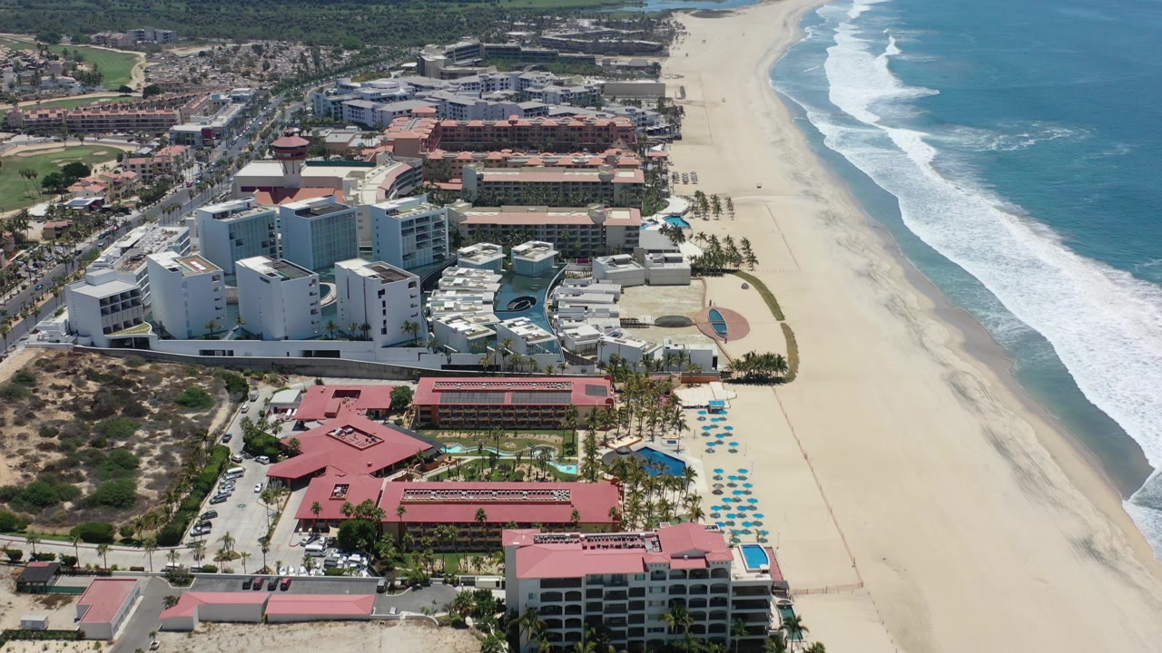 Drone view of beachside buildings, umbrellas, sand, and white ocean breaks.