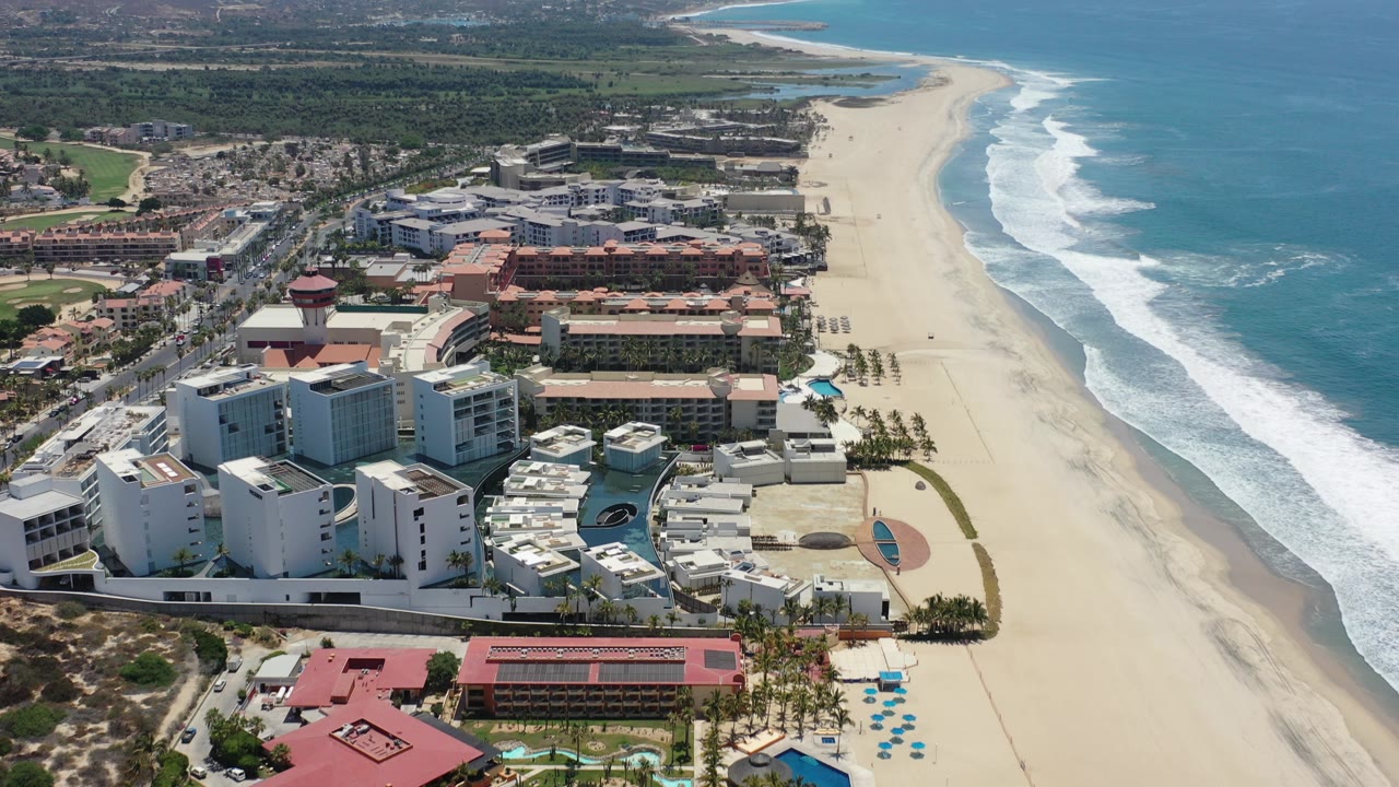 Aerial view over Los Cabos resort buildings, a broad sandy beach, and turquoise ocean surf, with no visible people in the frame.