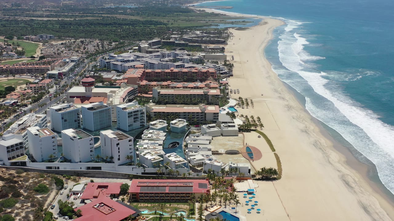 Aerial Los Cabos frame with white hotel blocks in the foreground, tan resort buildings behind them, and white waves rolling along the beach, with no visible people.