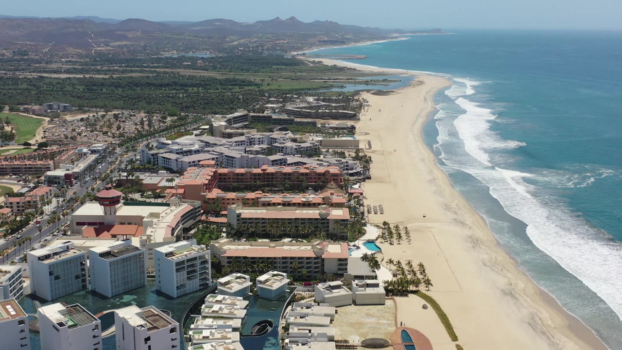 Aerial view along the Los Cabos coast, with foreground hotel roofs, a broad pale beach, parallel surf bands, turquoise water, inland greenery, distant hills, and no visible people.