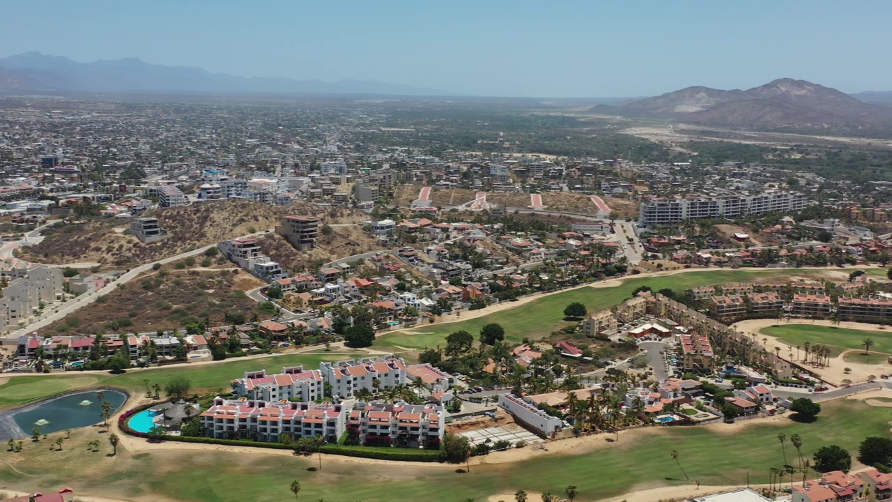 A high view over Los Cabos, Mexico centers green fairways and tan terrain with low-rise buildings, hillside roads, distant mountains, and no visible people.