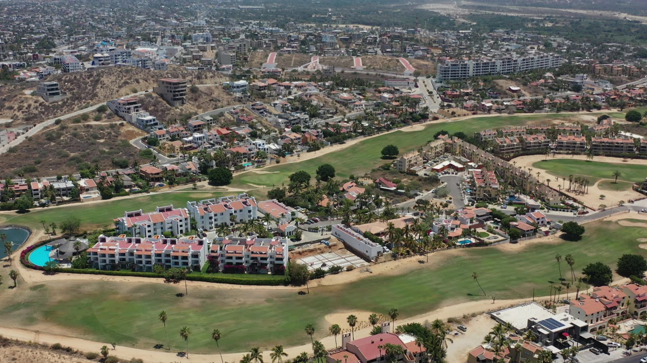 Opening aerial frame over Los Cabos, Mexico, showing green fields in front of red-roofed homes, swimming pools, roads, dry hills, and distant city blocks with no visible people.