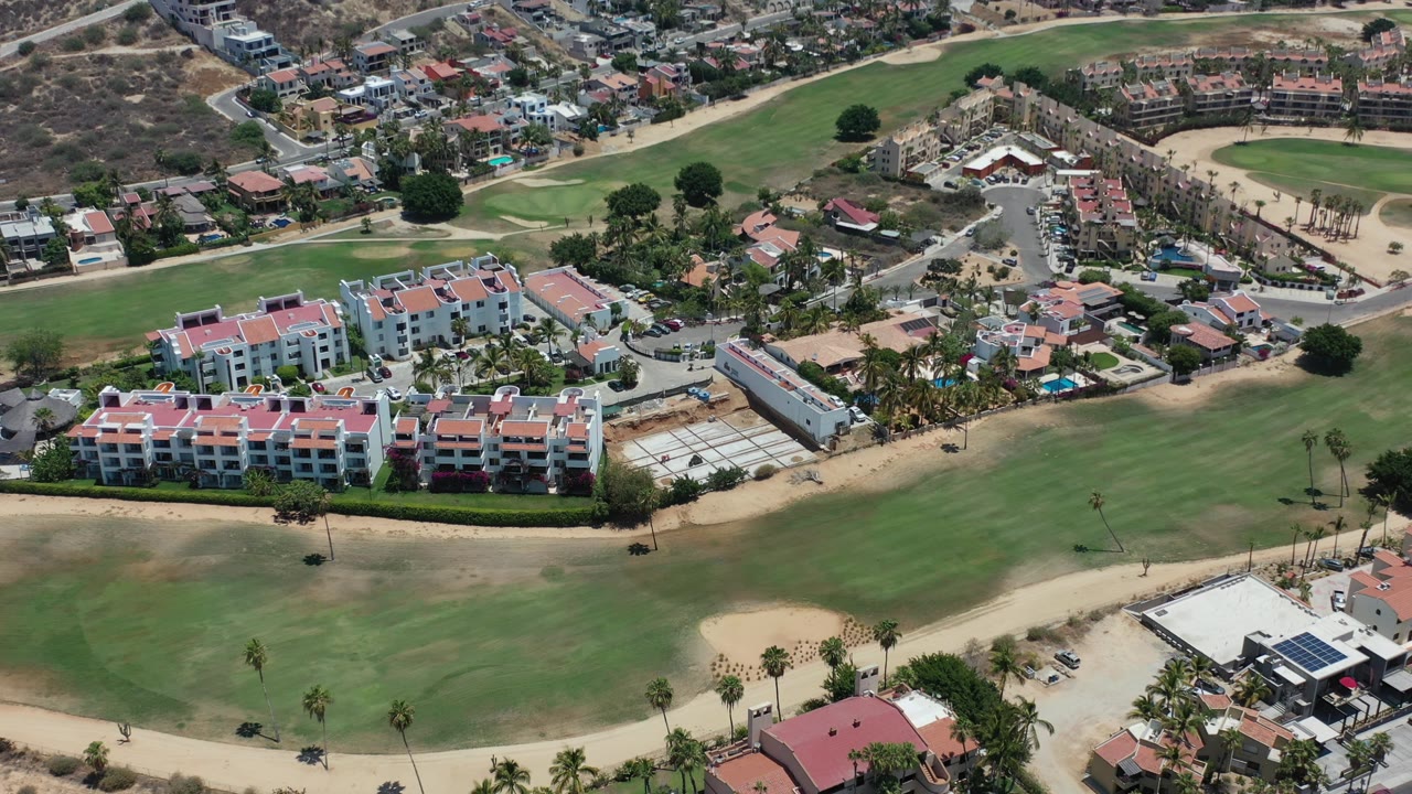 Aerial frame from Los Cabos, Mexico showing red-roofed residential buildings, sandy paths, green course lawns, roads, parked cars, and no visible people.