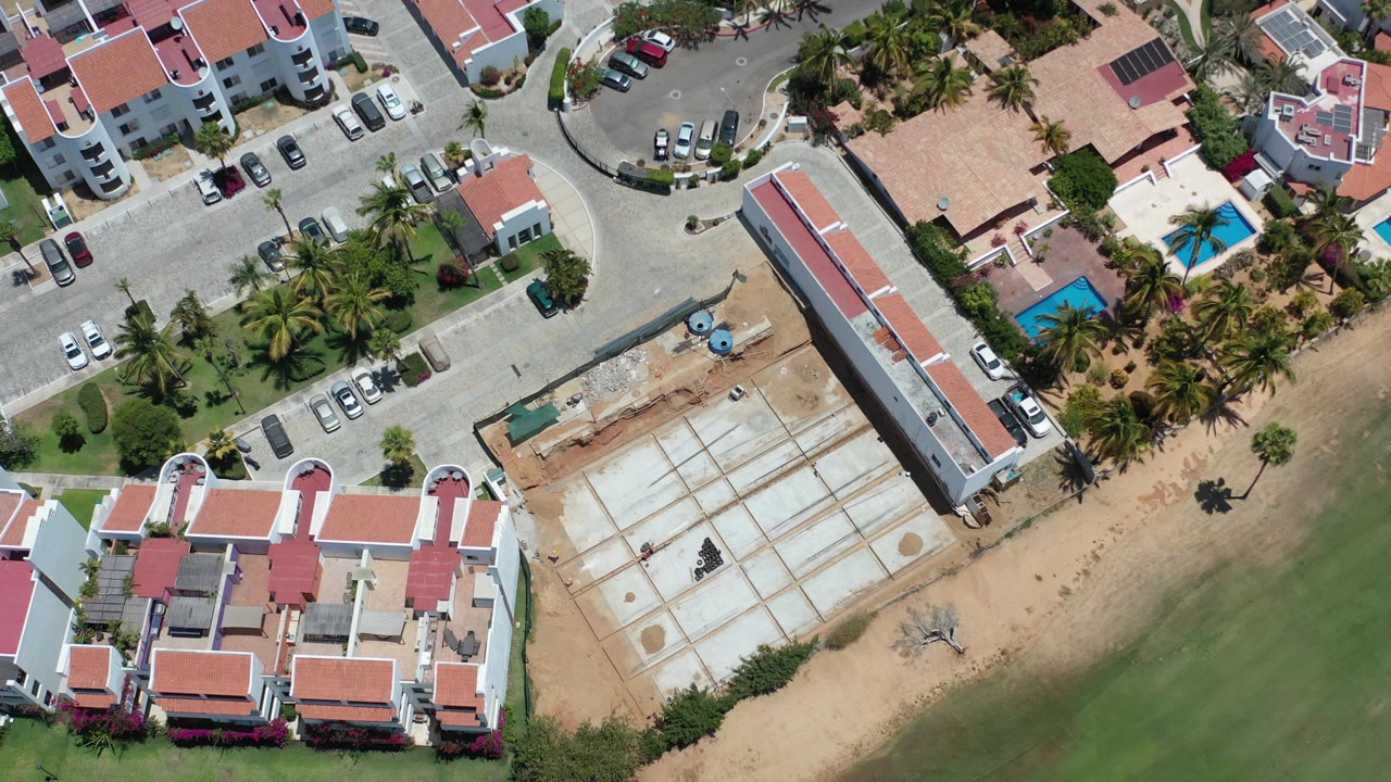 Top-down aerial frame in Los Cabos, Mexico, showing red-roofed buildings, a road curve, palm landscaping, a rectangular construction site, and green coastal water with no visible people.