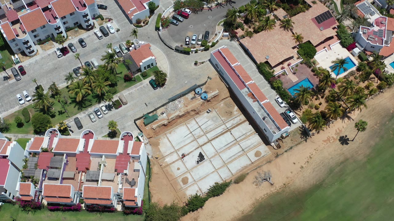 Los Cabos overhead view with the long red-roofed building near center right, parked cars along the road, palms beside a blue pool, and a pale construction grid next to green water, with no visible people.