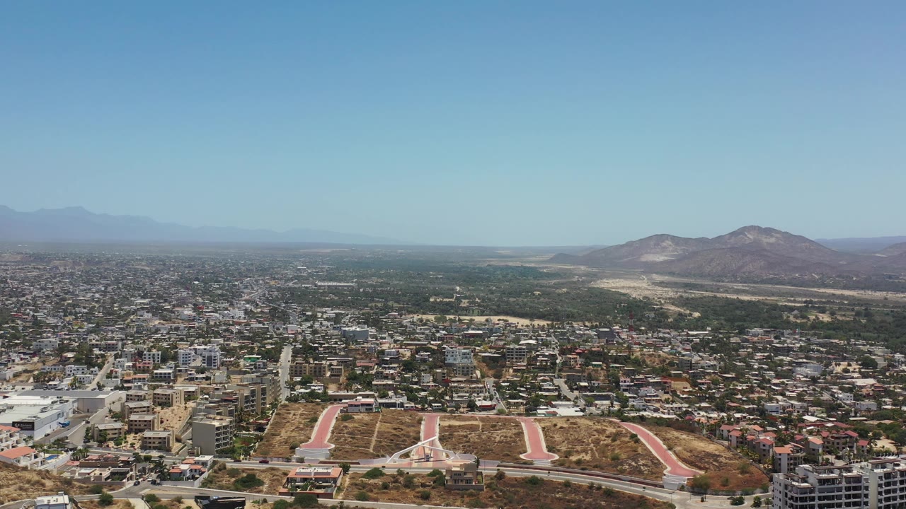 Early aerial frame of Los Cabos with compact buildings across the midground, dry brown lots in front, mountains on the horizon, and no visible people.