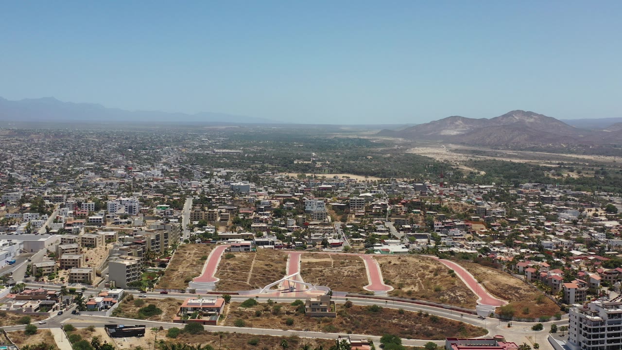 Opening aerial frame over Los Cabos, Mexico, showing dry undeveloped lots with pale pink paths in the foreground, dense city blocks beyond, distant mountains, blue sky, and no visible people.