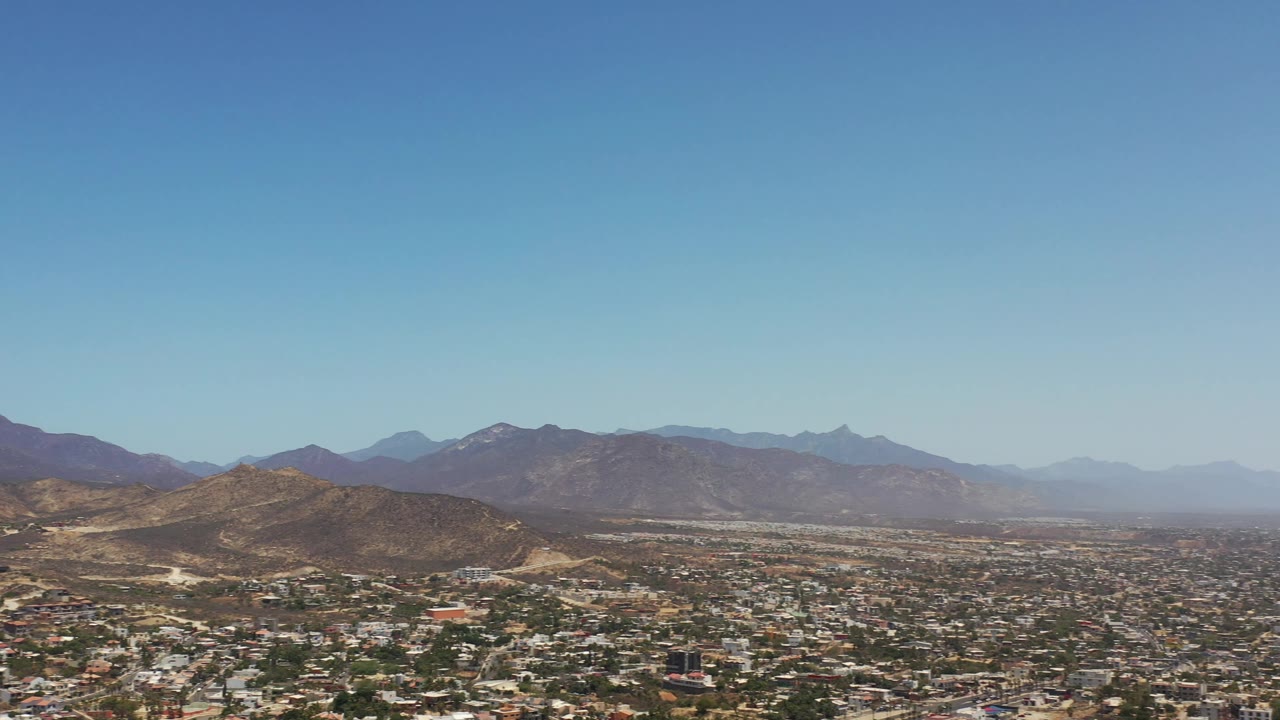 Los Cabos aerial frame with a rounded scrub-covered hill near center, low town blocks in the foreground, and layered mountains in the distance, with no visible people.
