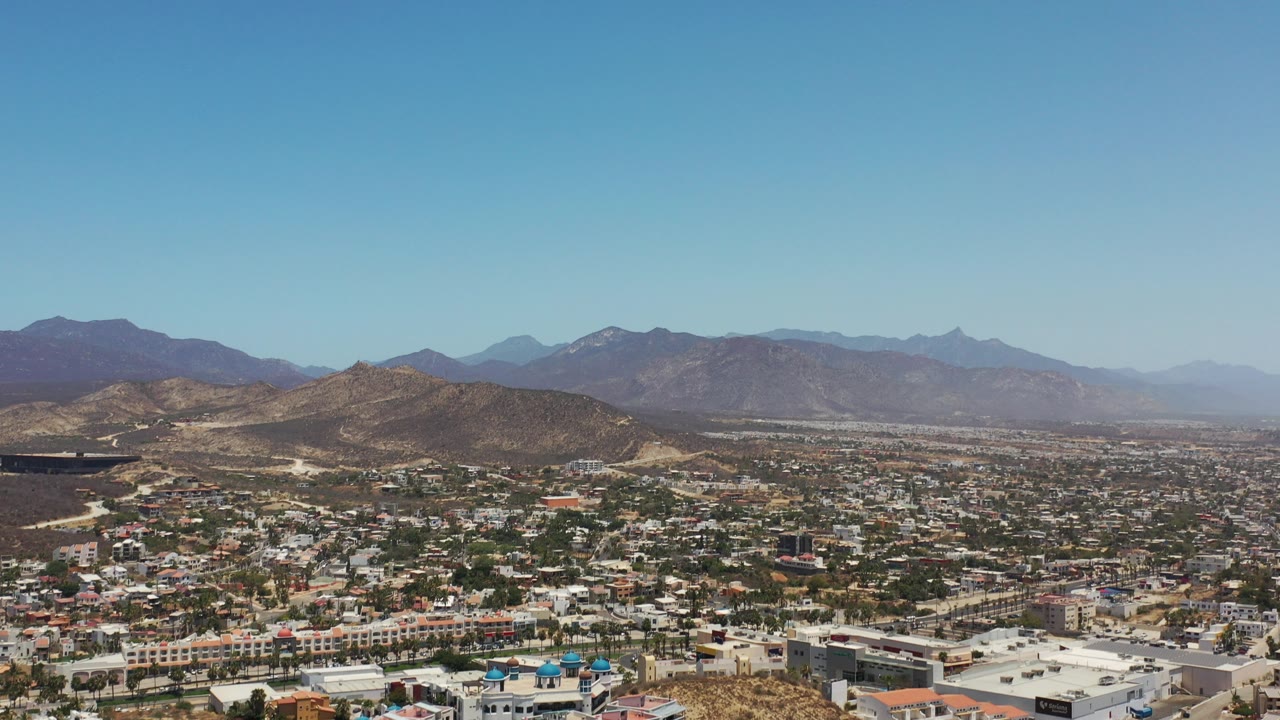 Wide aerial view of hillside buildings, trees, dry terrain, and distant mountains.