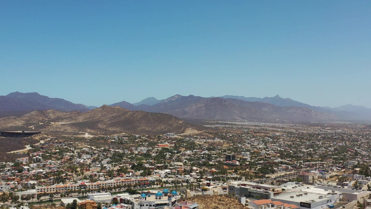 Los Cabos aerial view with a darker hillside at left, clustered buildings across the midground, hazy mountain ridges, and no visible people.