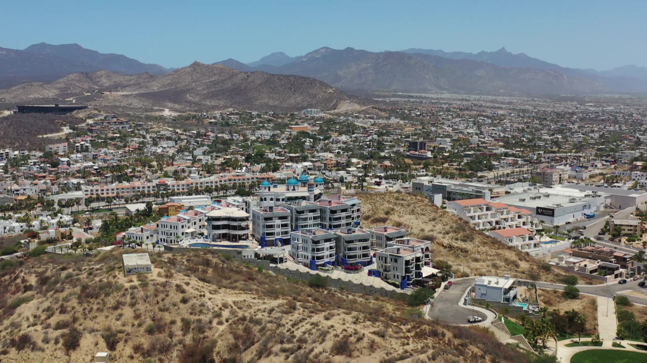 Early Los Cabos aerial frame with dry scrub-covered hills in front, white and gray building blocks in the middle distance, town streets beyond, and no visible people.
