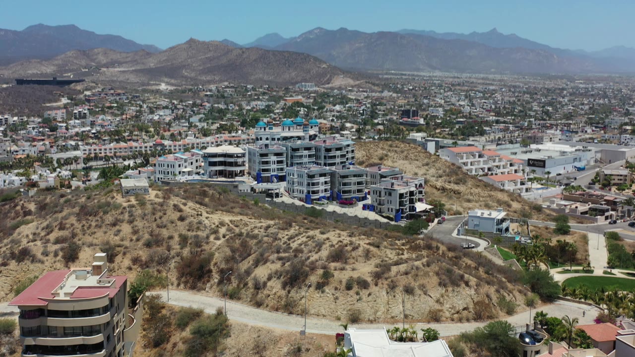 Second frame holds a wide Los Cabos hillside view with white multi-story homes in the midground, scrub-covered slopes, valley buildings, mountains, and no visible people.