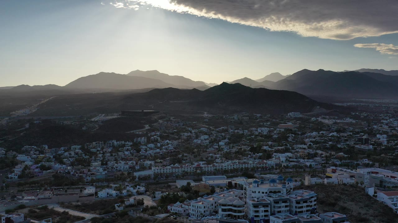 Los Cabos aerial view with clustered buildings in the lower frame, a dark hill across the middle, pale mountain layers on the horizon, bright sky, and no visible people.