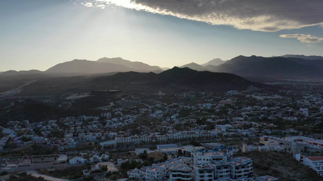 Aerial view in Los Cabos, Mexico, showing white residential buildings below dark hills, layered mountains, and sun rays breaking through cloud, with no visible people.